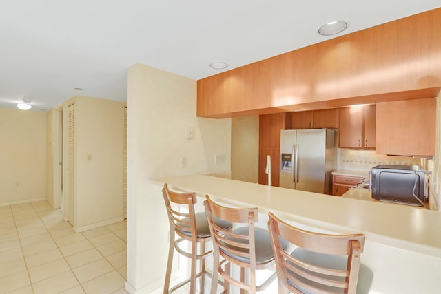 kitchen with light tile patterned flooring, stainless steel refrigerator with ice dispenser, decorative backsplash, a kitchen bar, and kitchen peninsula