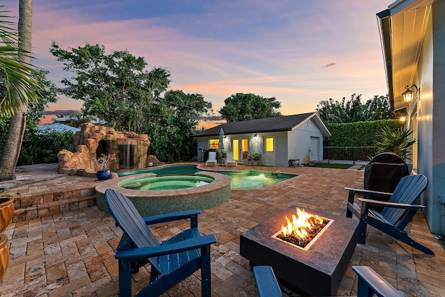 pool at dusk featuring an in ground hot tub, an outdoor structure, a patio area, and a fire pit