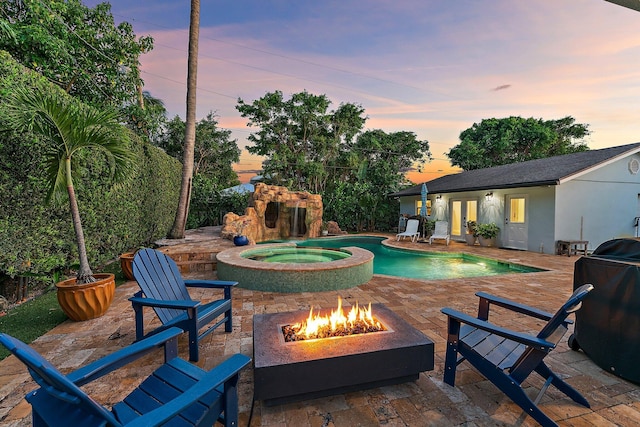 pool at dusk featuring an in ground hot tub, an outdoor structure, a patio area, and a fire pit