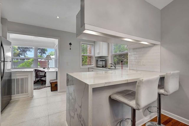 kitchen featuring a breakfast bar area, kitchen peninsula, white cabinets, light stone countertops, and backsplash