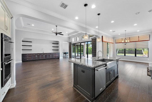 kitchen with decorative light fixtures, ornamental molding, an island with sink, light stone countertops, and dark wood-type flooring