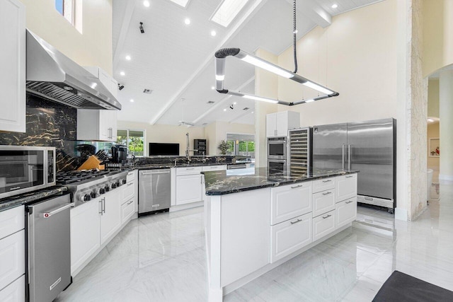 kitchen with dark stone countertops, stainless steel appliances, wall chimney range hood, and high vaulted ceiling