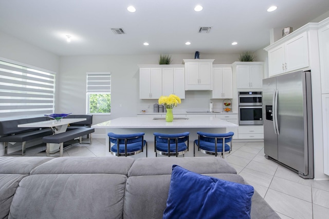 kitchen featuring a kitchen island with sink, white cabinets, light tile patterned flooring, stainless steel appliances, and a breakfast bar