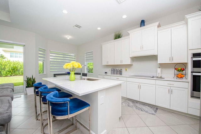 kitchen featuring a center island with sink, double oven, white cabinets, light tile patterned floors, and sink