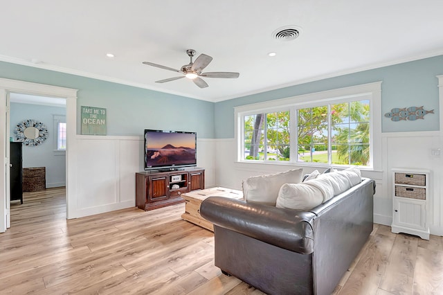 living area featuring a ceiling fan, wainscoting, light wood-style flooring, visible vents, and ornamental molding