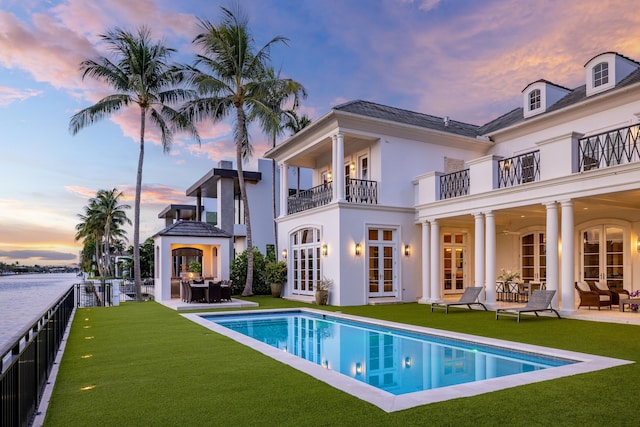 back house at dusk featuring french doors, a balcony, a patio, a fenced in pool, and a lawn