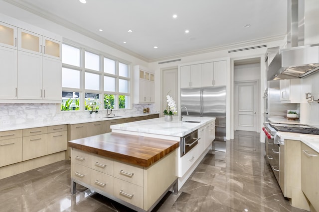 kitchen featuring wooden counters, backsplash, crown molding, wall chimney range hood, and an island with sink