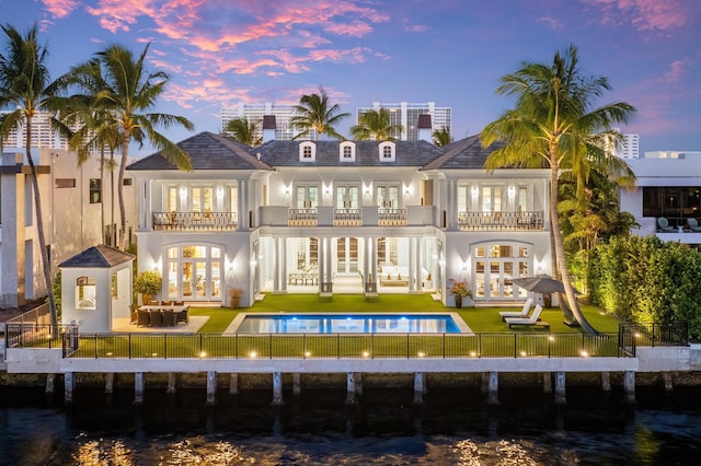 back house at dusk with a lawn, french doors, a balcony, a patio area, and a water view