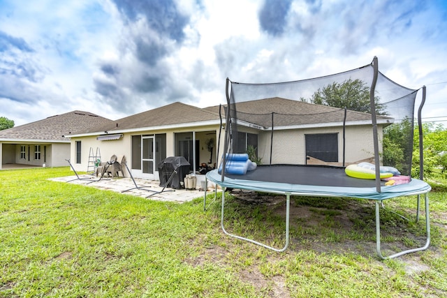 back of house featuring a trampoline, roof with shingles, a yard, stucco siding, and a patio area