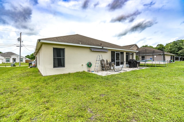 rear view of property with a trampoline, roof with shingles, stucco siding, a lawn, and a patio area