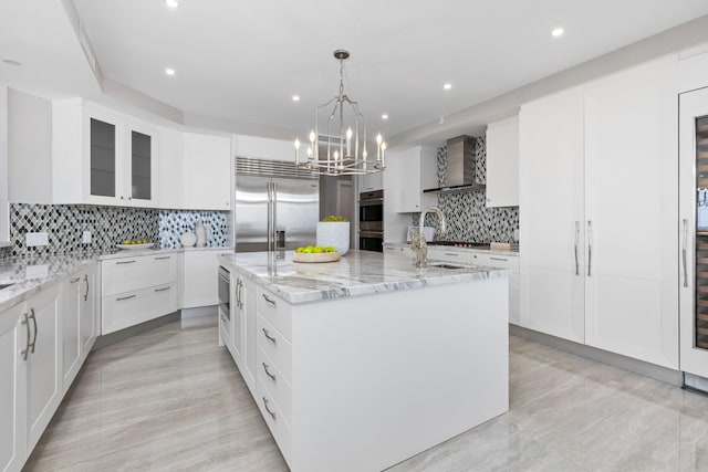 kitchen with wall chimney range hood, a kitchen island with sink, light stone countertops, appliances with stainless steel finishes, and decorative backsplash