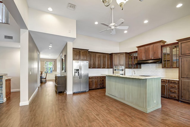 kitchen featuring ceiling fan, a kitchen island with sink, stainless steel fridge, hardwood / wood-style floors, and decorative backsplash