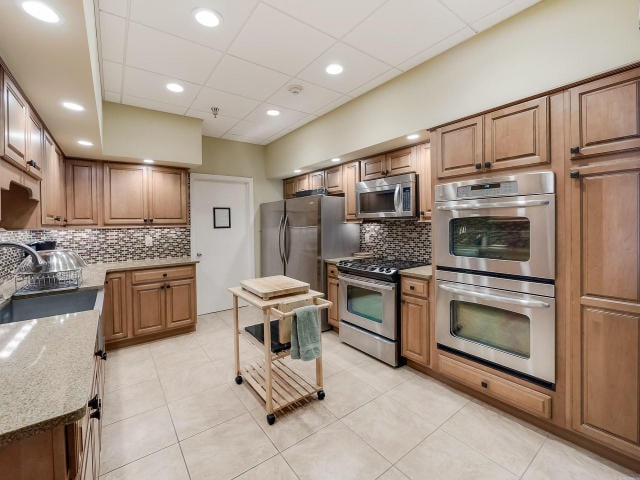 kitchen with light tile patterned floors, appliances with stainless steel finishes, tasteful backsplash, light stone countertops, and a drop ceiling