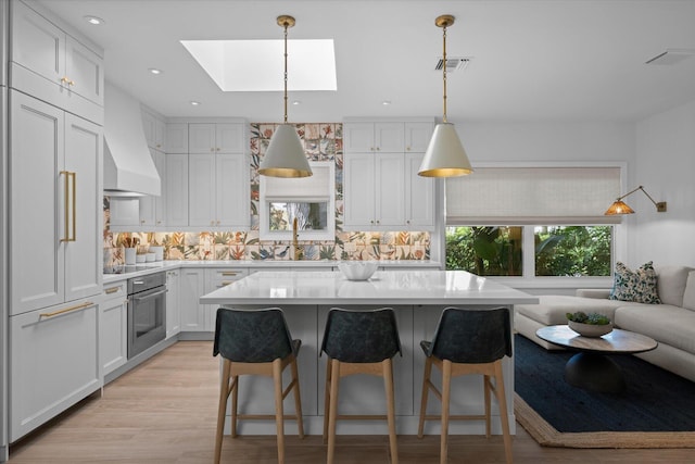 kitchen featuring light wood-type flooring, a skylight, stainless steel oven, decorative backsplash, and custom range hood