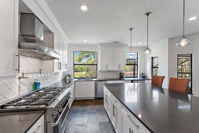 kitchen featuring appliances with stainless steel finishes, wall chimney exhaust hood, a healthy amount of sunlight, white cabinetry, and hanging light fixtures