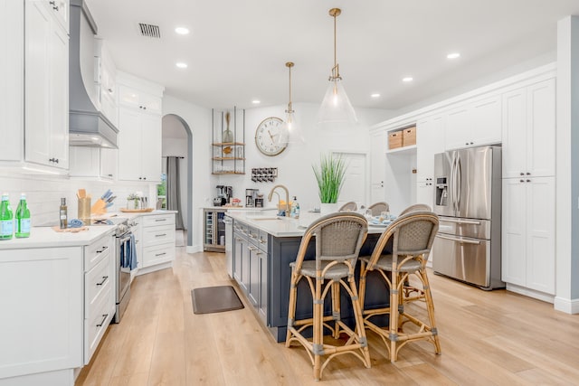 kitchen featuring stainless steel appliances, an island with sink, white cabinets, custom range hood, and light wood-type flooring