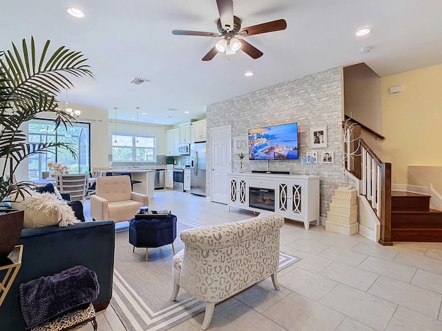 living room with a stone fireplace, ceiling fan with notable chandelier, light tile patterned floors, and sink