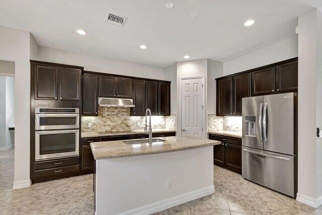 kitchen featuring appliances with stainless steel finishes, an island with sink, sink, dark brown cabinetry, and light stone counters