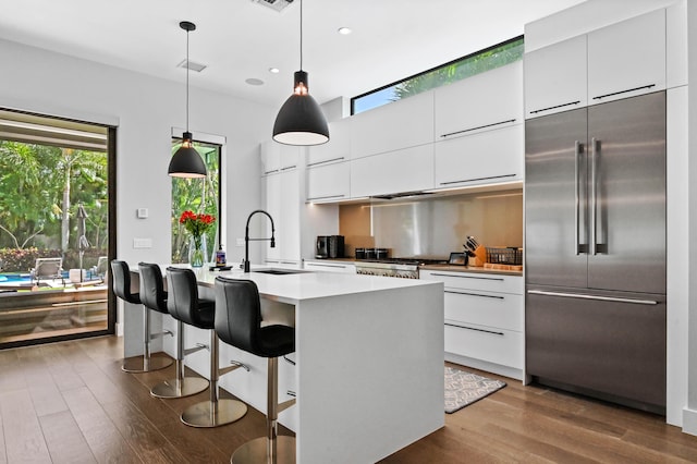 kitchen with built in fridge, pendant lighting, light countertops, a kitchen island with sink, and white cabinetry