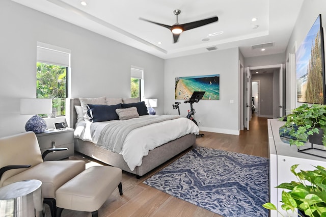 bedroom with ceiling fan, a raised ceiling, wood-type flooring, and multiple windows