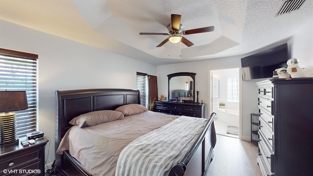 bedroom with a textured ceiling, ensuite bathroom, visible vents, light wood finished floors, and a tray ceiling