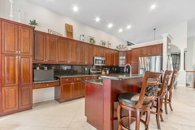 kitchen featuring backsplash, stainless steel appliances, a center island with sink, light tile patterned floors, and dark stone counters