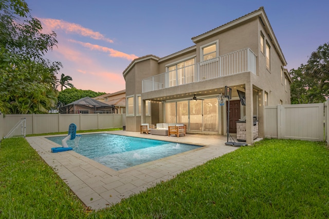 back house at dusk with a lawn, a fenced in pool, a balcony, ceiling fan, and a patio