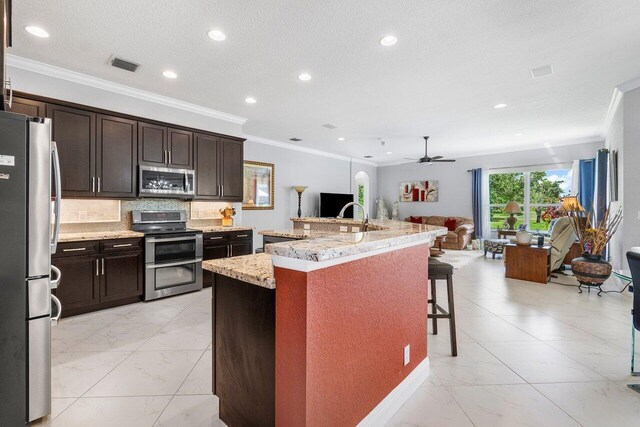 kitchen with a kitchen bar, an island with sink, stainless steel appliances, ceiling fan, and light stone counters