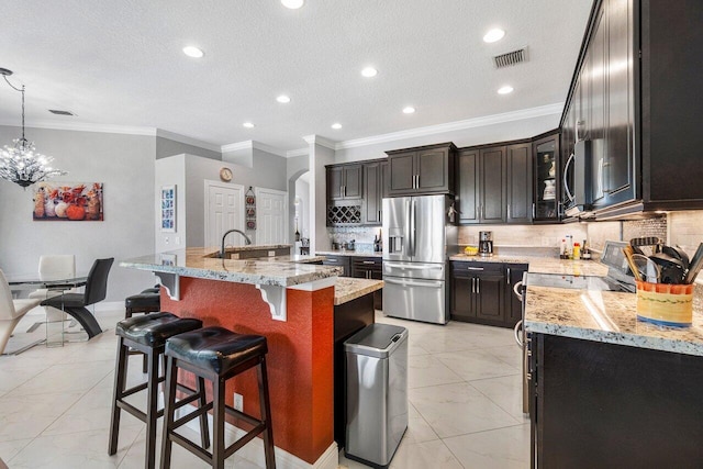 kitchen featuring ornamental molding, stainless steel appliances, decorative light fixtures, light stone counters, and a center island with sink