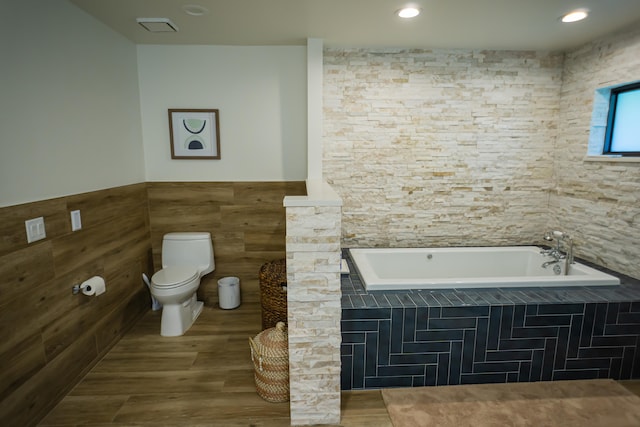 bathroom featuring a relaxing tiled tub, toilet, tile walls, and hardwood / wood-style flooring