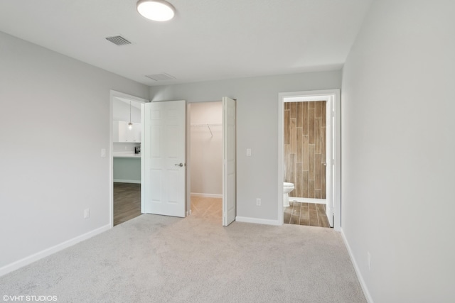 unfurnished bedroom featuring a spacious closet, a closet, light colored carpet, and ensuite bath