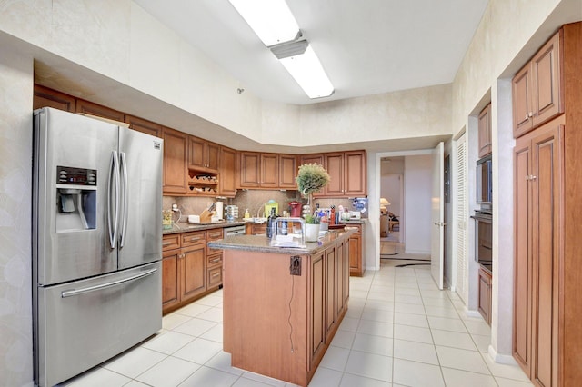 kitchen featuring a center island with sink, light tile patterned floors, stainless steel appliances, tasteful backsplash, and brown cabinetry