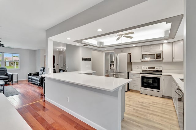 kitchen featuring appliances with stainless steel finishes, a raised ceiling, light hardwood / wood-style floors, kitchen peninsula, and ceiling fan