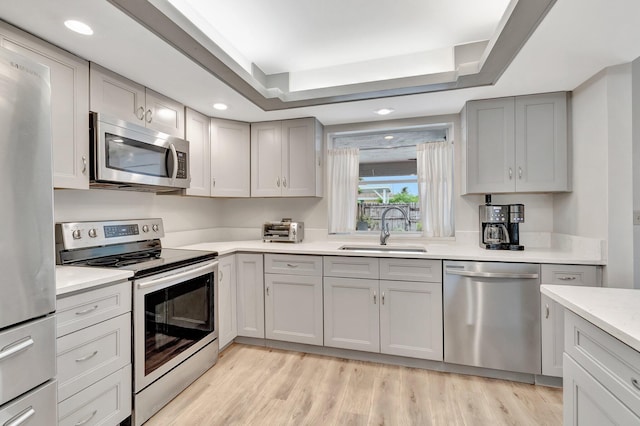 kitchen featuring stainless steel appliances, sink, gray cabinetry, a tray ceiling, and light wood-type flooring