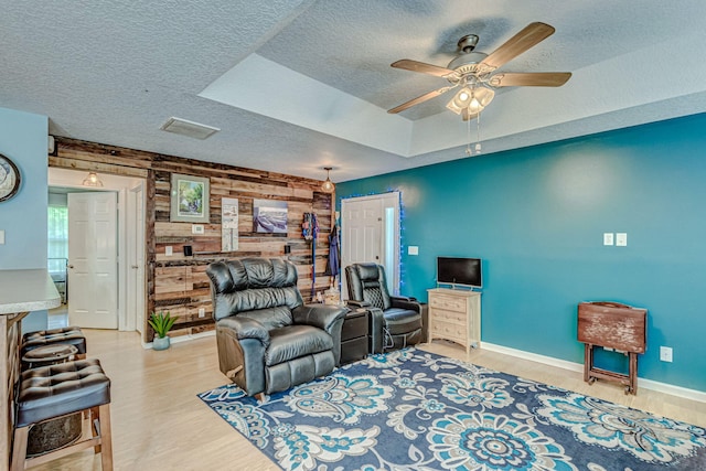 living room with ceiling fan, light wood-type flooring, wooden walls, a tray ceiling, and a textured ceiling