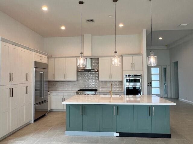 kitchen with wall chimney range hood, a center island with sink, white cabinets, pendant lighting, and appliances with stainless steel finishes