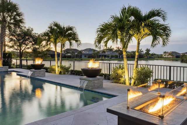 pool at dusk with pool water feature, a water view, an outdoor fire pit, and a patio area