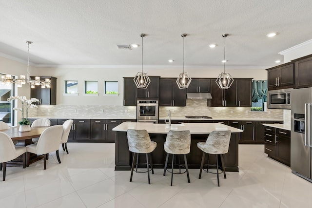 kitchen with dark brown cabinetry, stainless steel appliances, hanging light fixtures, crown molding, and a center island with sink