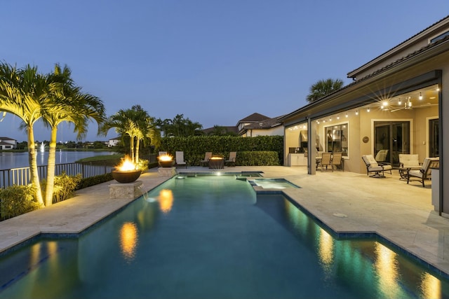 pool at dusk with an in ground hot tub, a water view, and a patio area