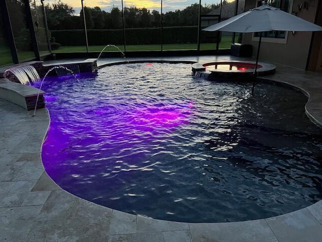 pool at dusk with a patio, an in ground hot tub, pool water feature, and glass enclosure