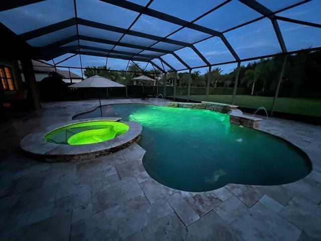 pool at dusk featuring a patio, an in ground hot tub, pool water feature, and glass enclosure