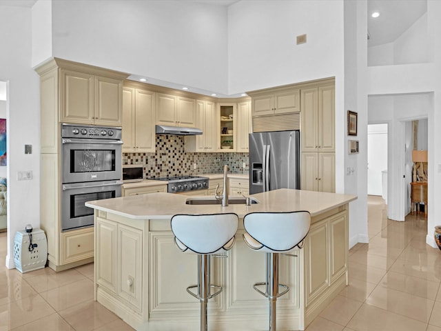 kitchen featuring light tile patterned flooring, a kitchen bar, appliances with stainless steel finishes, an island with sink, and a towering ceiling