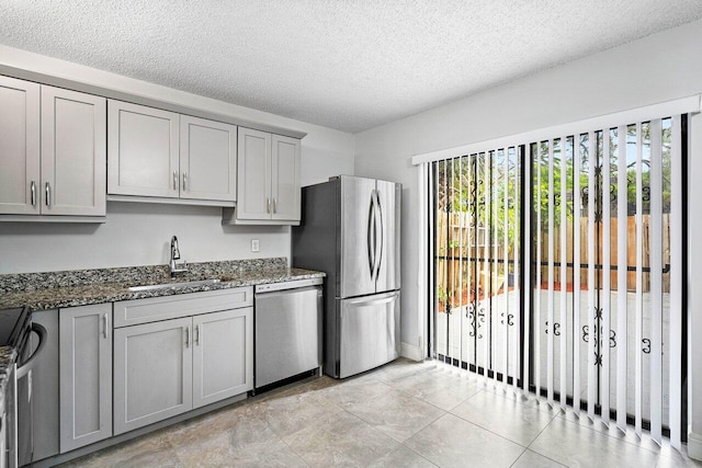 kitchen with stainless steel appliances, gray cabinetry, a sink, a textured ceiling, and dark stone countertops