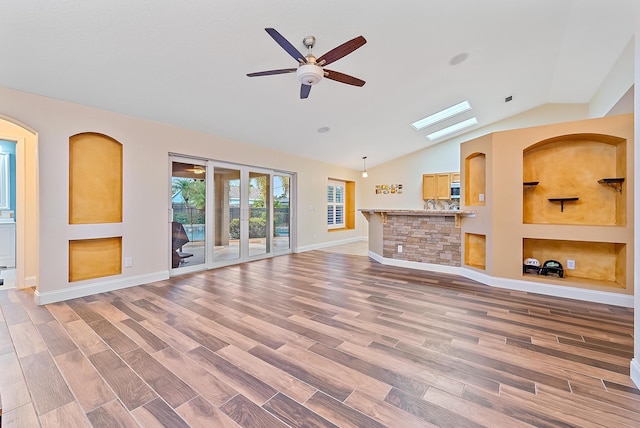 unfurnished living room with vaulted ceiling with skylight, ceiling fan, built in shelves, and light wood-type flooring