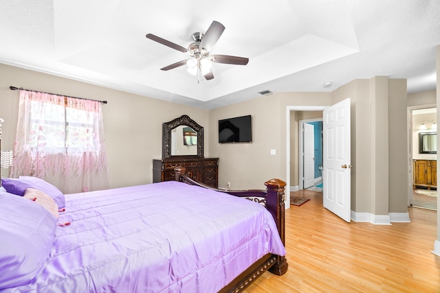 bedroom with ceiling fan, light wood-type flooring, ensuite bathroom, and a tray ceiling