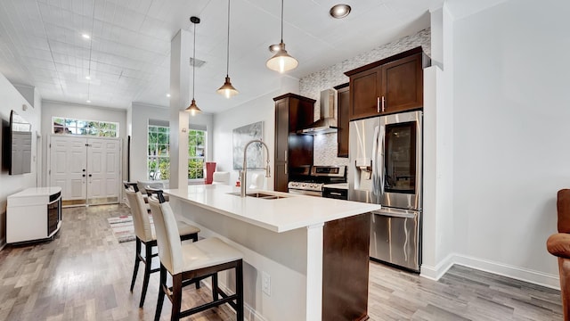 kitchen featuring appliances with stainless steel finishes, light hardwood / wood-style floors, wall chimney exhaust hood, a center island with sink, and pendant lighting