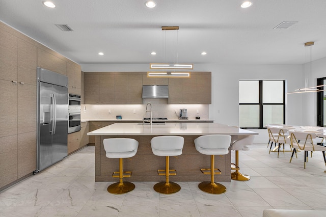 kitchen with backsplash, appliances with stainless steel finishes, exhaust hood, hanging light fixtures, and a center island with sink