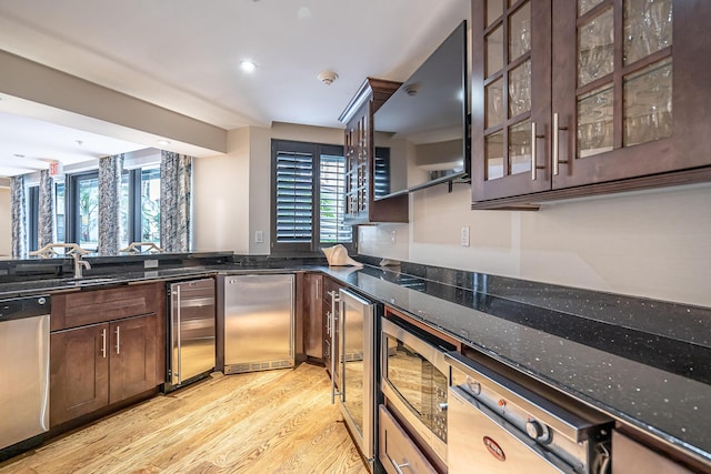 kitchen featuring light wood-style flooring, dark brown cabinetry, beverage cooler, stainless steel appliances, and dark stone countertops