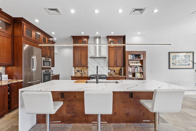 kitchen featuring wall chimney exhaust hood, appliances with stainless steel finishes, tile patterned floors, decorative backsplash, and a large island with sink