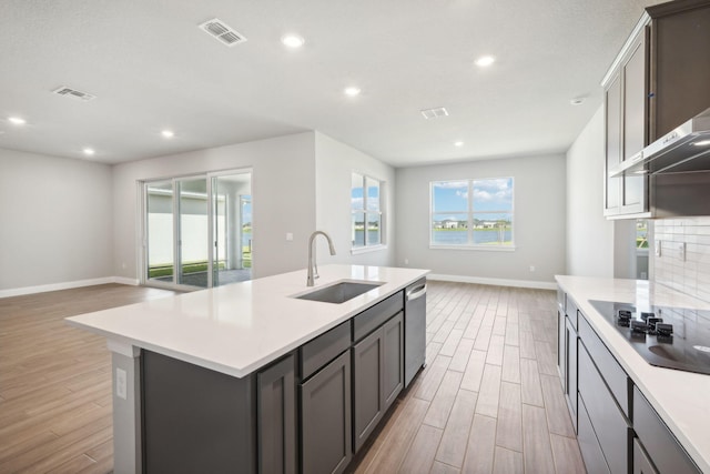 kitchen with black electric stovetop, sink, a kitchen island with sink, stainless steel dishwasher, and light hardwood / wood-style flooring